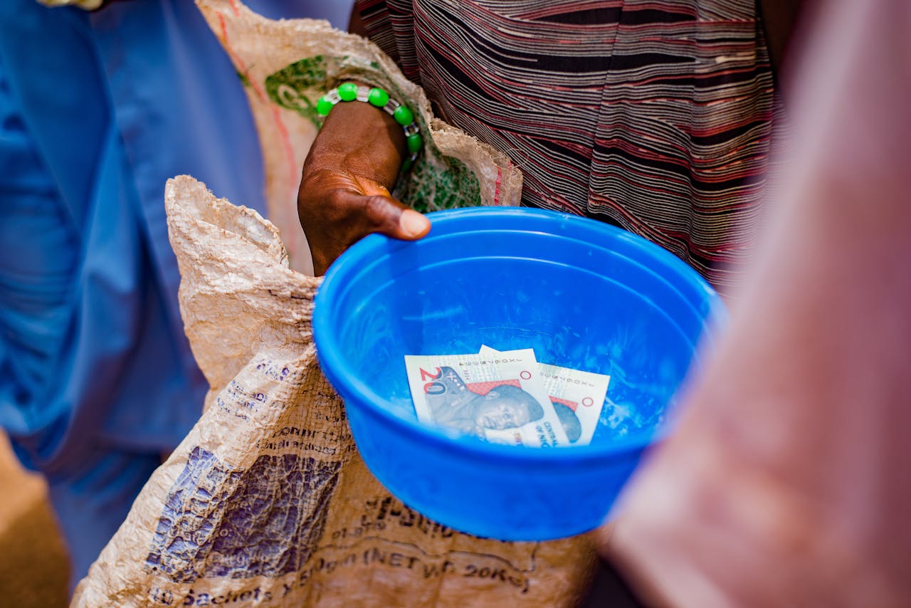 A close-up of a hand holding a blue bowl containing currency notes outdoors.