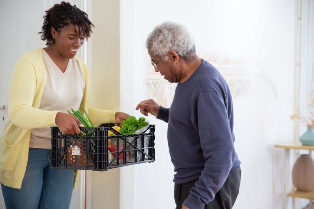 pexels photo 7551597 A young adult woman helps a senior man by carrying a crate of fresh vegetables indoors, symbolizing care and support.